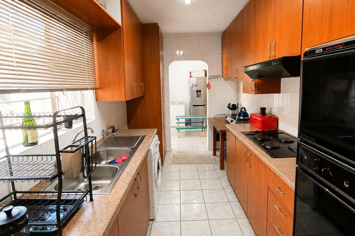 Functional galley-style kitchen featuring wood-look cabinetry, built-in electric oven and hob, double sink, and tiled flooring with direct access to a secondary utility area.