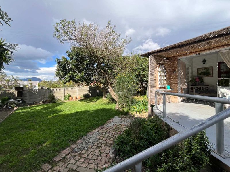 A rear garden view featuring a lawn, cobblestone path, and a covered patio area with a metal railing and thatched roof detail.