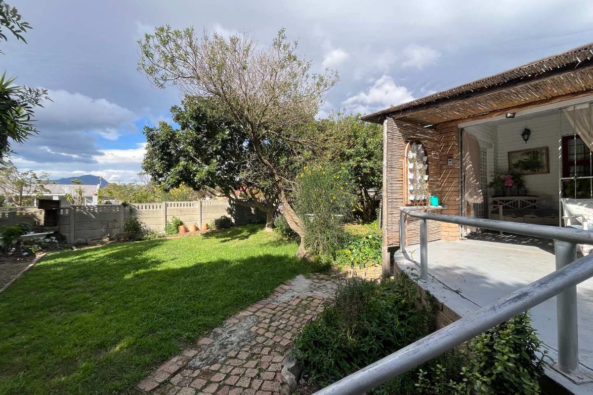 A rear garden view featuring a lawn, cobblestone path, and a covered patio area with a metal railing and thatched roof detail.