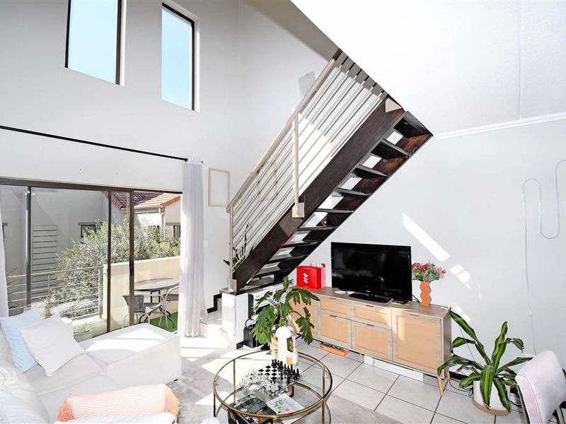 Bright double-volume living area featuring a modern staircase with metal balustrade, tiled flooring, and large sliding doors leading to an outdoor balcony.