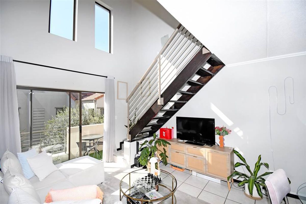 Bright double-volume living area featuring a modern staircase with metal balustrade, tiled flooring, and large sliding doors leading to an outdoor balcony.