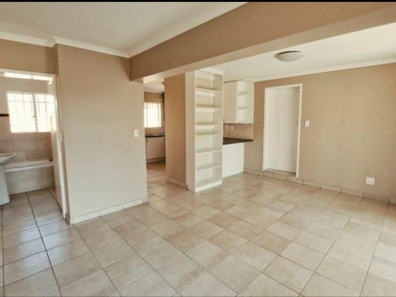An open-plan living area featuring neutral ceramic floor tiles, built-in shelving, and access to an adjacent bathroom and kitchen space.