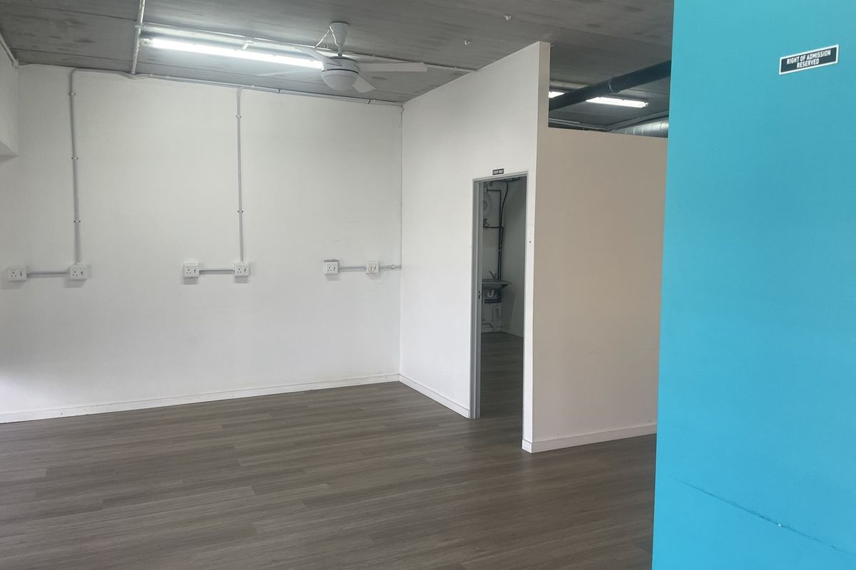 An open-plan retail space featuring laminate flooring, exposed ceilings, surface-mounted electrical conduits, and a separate utility area.