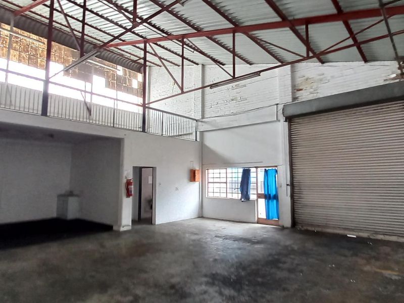 Spacious industrial warehouse unit featuring a roller shutter door, mezzanine level, exposed steel roof trusses, and concrete flooring, suitable for various commercial or storage uses.