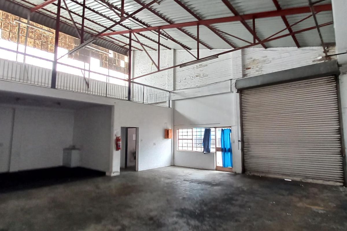 Spacious industrial warehouse unit featuring a roller shutter door, mezzanine level, exposed steel roof trusses, and concrete flooring, suitable for various commercial or storage uses.