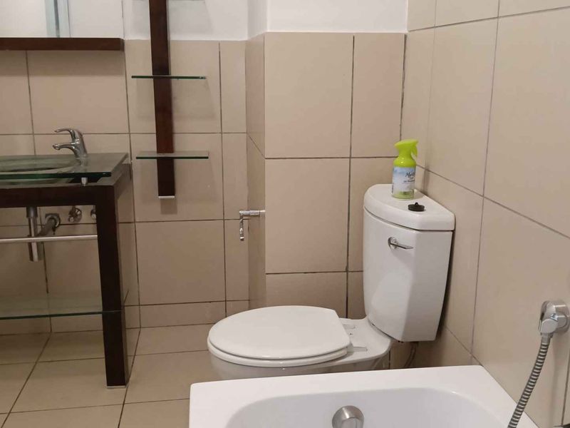 A functional bathroom featuring a glass basin vanity, toilet, and wall-mounted shower head with neutral wall and floor tiling.