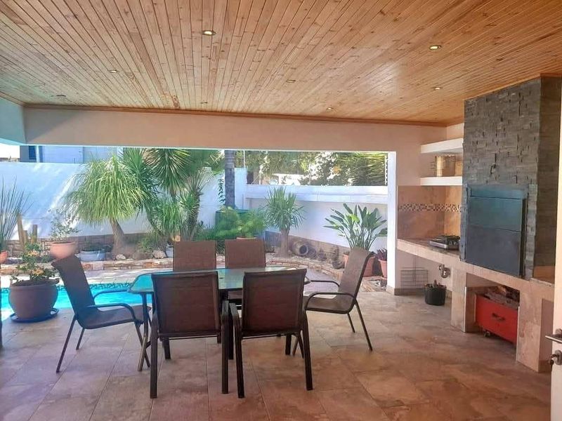 A covered outdoor patio featuring a timber ceiling, recessed lighting, and a built-in braai area, overlooking a private swimming pool and garden.