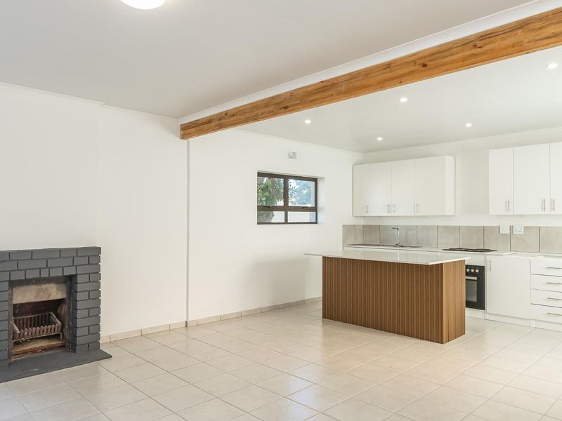 Open-plan living and kitchen area featuring a brick fireplace, exposed timber beam, modern kitchen island, and neutral tiled flooring throughout.