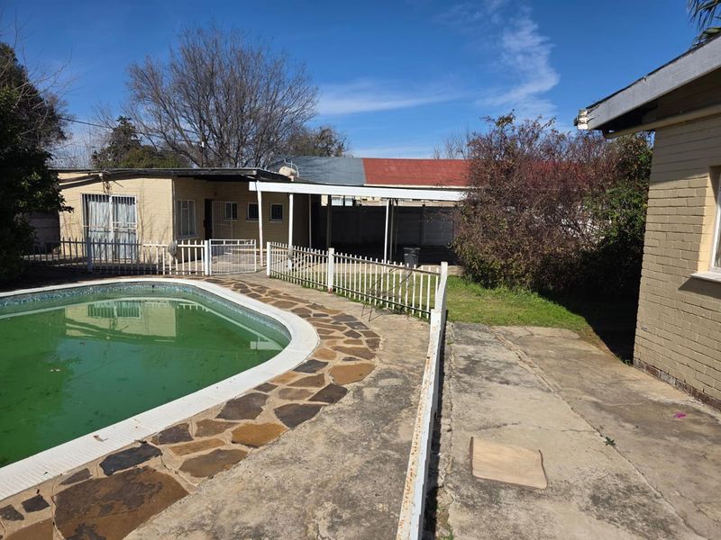 Outdoor pool area featuring a paved surround, metal safety fencing, and a covered patio space, requiring maintenance and pool restoration.