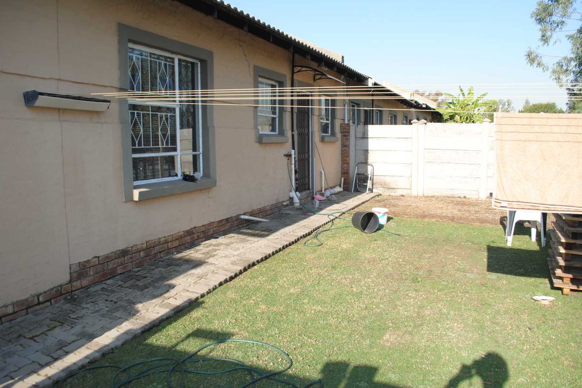 Rear exterior view of the townhouse featuring a paved walkway, small lawn area, security gates, and perimeter walling.
