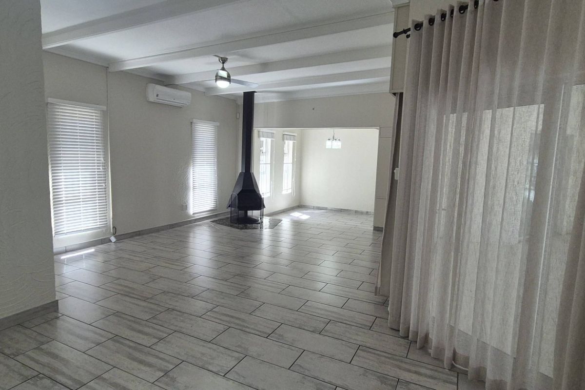 Spacious, light-filled living area featuring modern tiled flooring, exposed ceiling beams, a central freestanding fireplace, and split-system air conditioning.