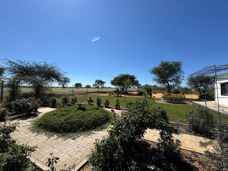 A well-maintained farm garden featuring a paved walkway, established rose bushes, a raised garden bed, and a metal trellis, overlooking open rural landscape.