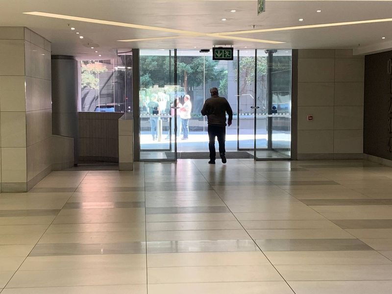 A spacious and modern office building entrance lobby featuring tiled flooring, recessed ceiling lighting, and large glass sliding doors.