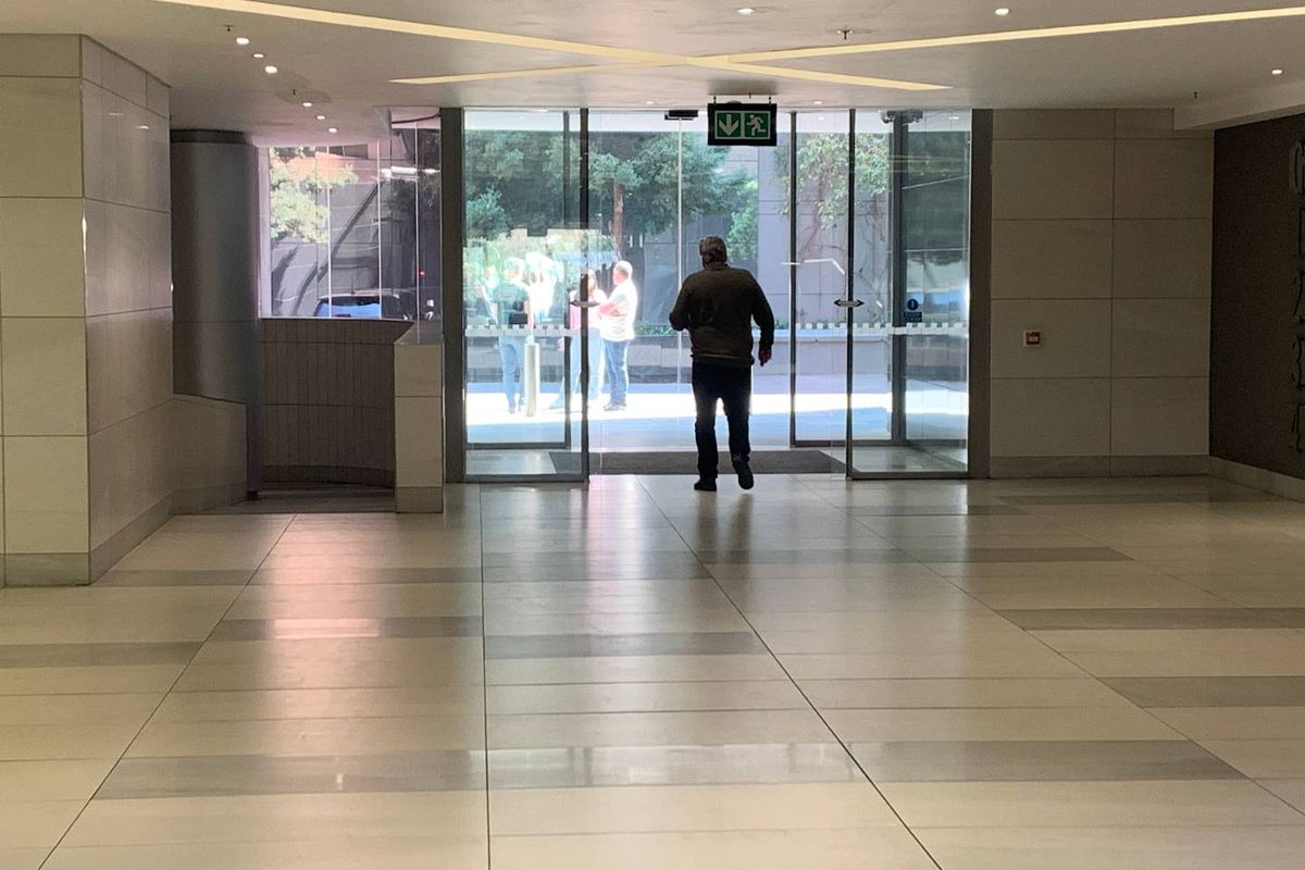 A spacious and modern office building entrance lobby featuring tiled flooring, recessed ceiling lighting, and large glass sliding doors.