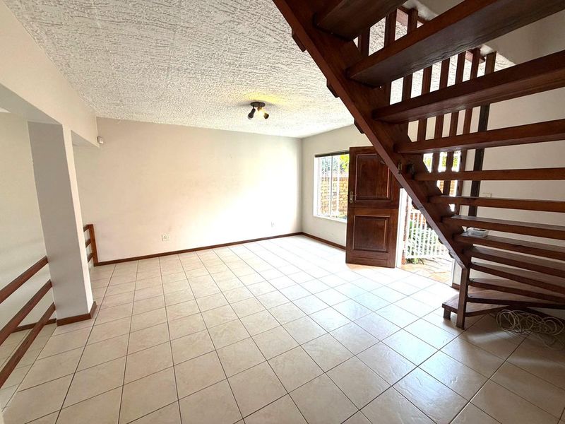 An open-plan living area featuring tiled flooring, a prominent wooden staircase, and direct access to the exterior through a timber entrance door.