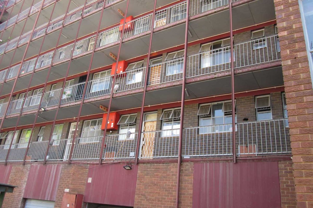Exterior view of a multi-story apartment building featuring brick facade, metal balcony railings, and security gates on multiple levels.