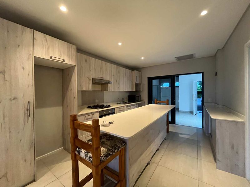 Modern kitchen featuring light wood-grain cabinetry, a central island with stone countertops, integrated gas hob, and tiled flooring with direct access to the exterior.