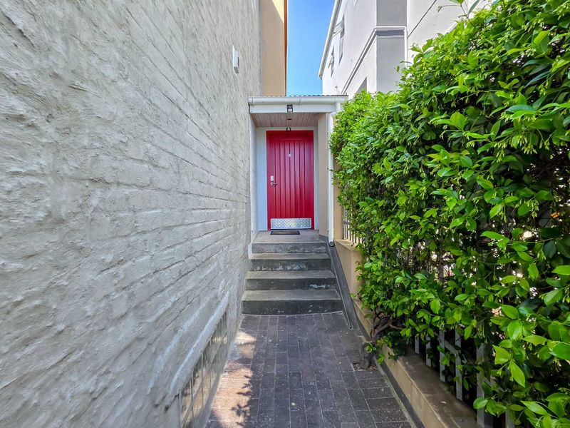 A side access path featuring a red entrance door, concrete steps, and a paved walkway bordered by a painted brick wall and established hedging.