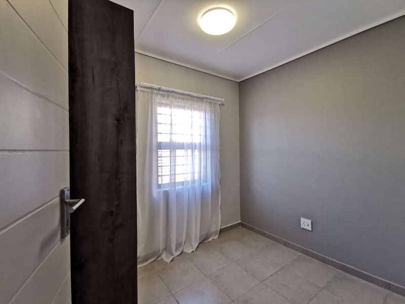 A clean, empty bedroom featuring neutral grey walls, tiled flooring, and a window fitted with security bars.