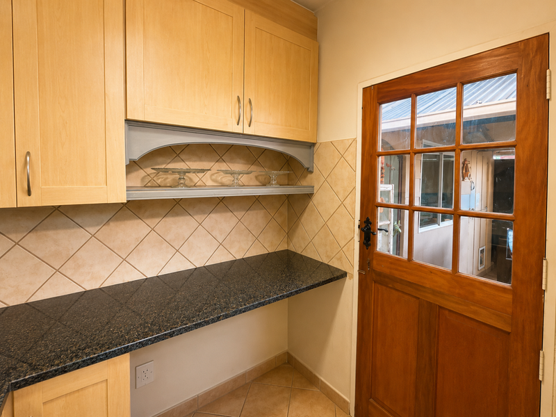 A functional scullery area featuring light wood cabinetry, dark granite countertops, tiled backsplash, and a timber-framed glass door leading to the exterior.