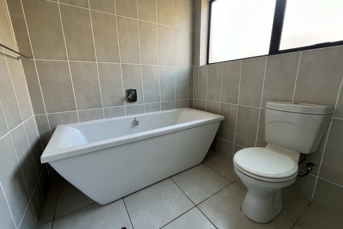 A modern bathroom featuring a freestanding bathtub, a standard toilet, and neutral floor-to-ceiling wall tiling with a large window for natural light.