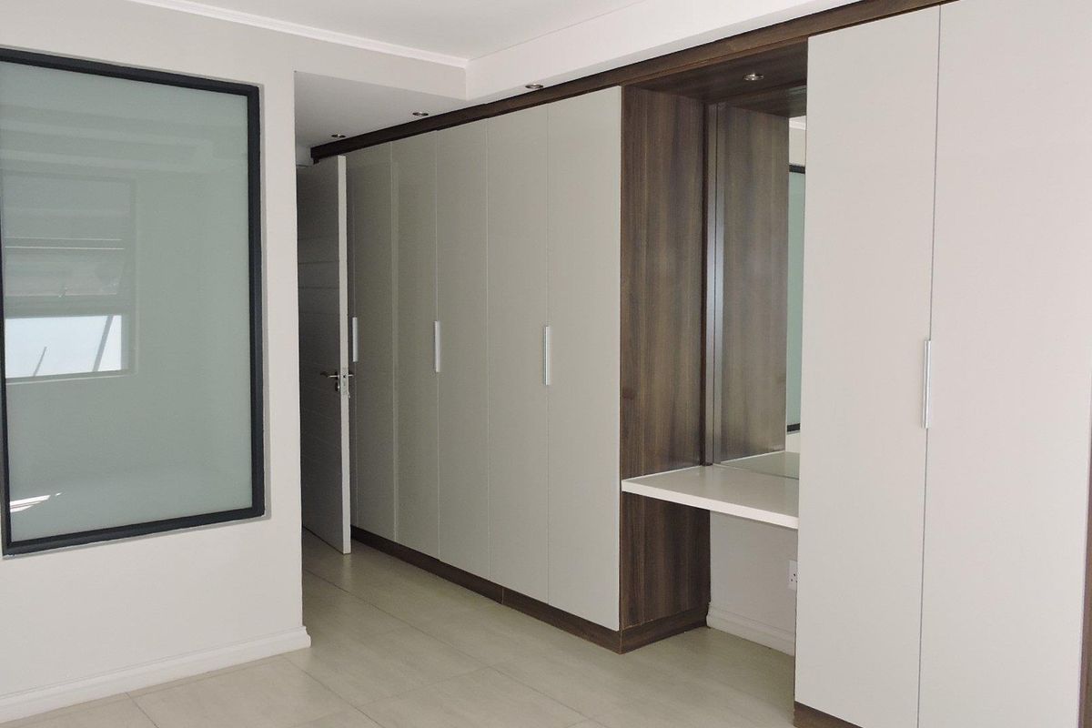 A modern bedroom featuring extensive built-in cabinetry with an integrated vanity, neutral tiled flooring, and a stylish frosted glass feature panel.
