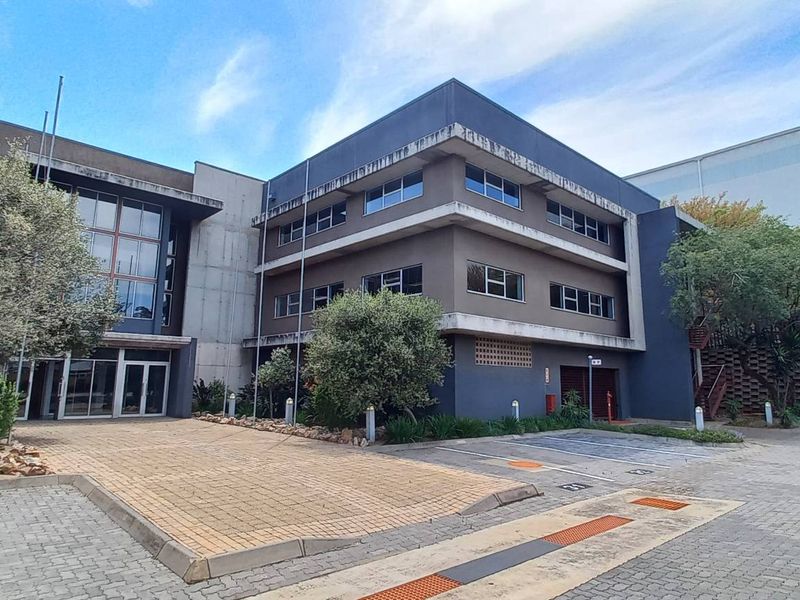 A multi-story office building exterior featuring a paved driveway, landscaped garden, and modern architectural facade with large windows.