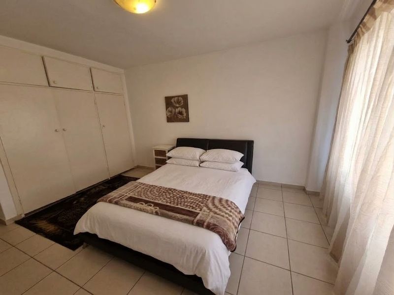 A functional bedroom featuring tiled flooring, built-in cupboards for storage, and large windows with curtain rails.