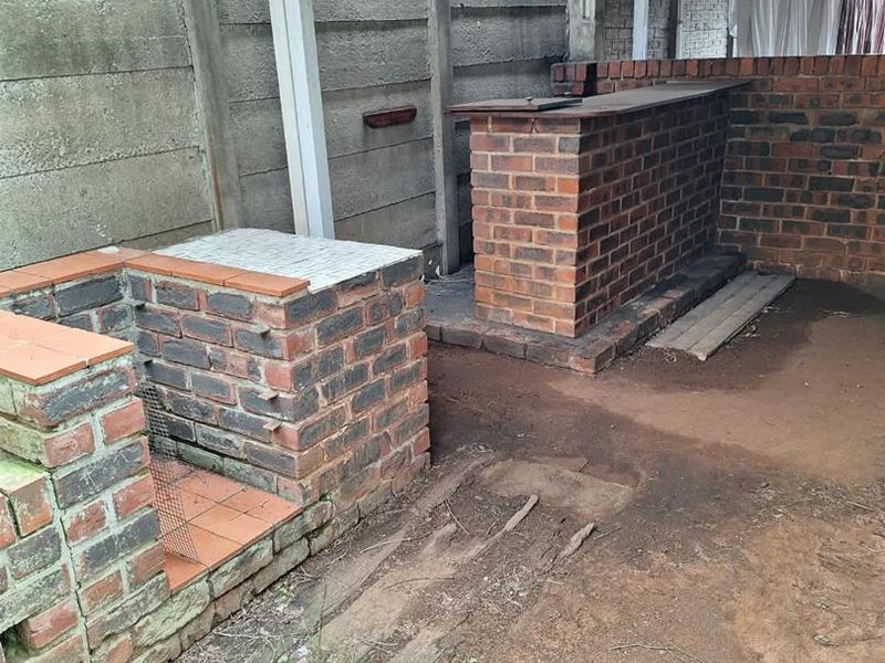 Outdoor covered patio area featuring a built-in brick braai and bar counter, set against a precast boundary wall with an unfinished dirt floor.