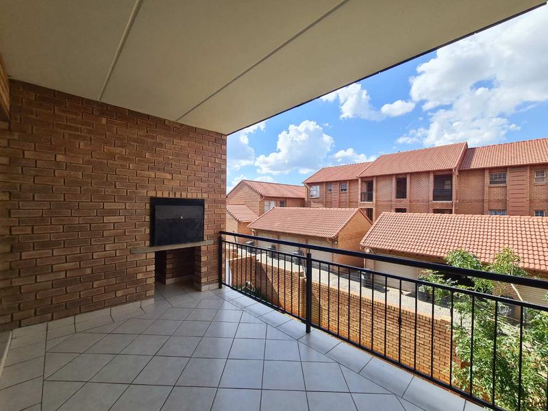 A spacious covered balcony featuring tiled flooring, a built-in braai, and a metal balustrade, offering an elevated view of the surrounding apartment complex.