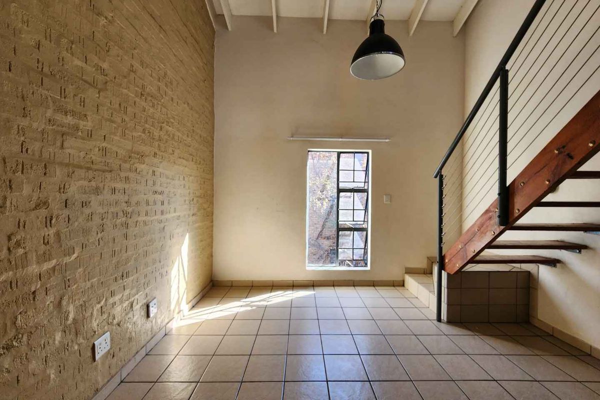 An empty living area featuring an exposed brick wall, tiled flooring, and a staircase with a metal railing, offering a unique industrial aesthetic.