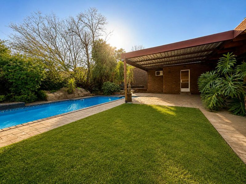 A well-maintained outdoor area featuring a swimming pool, a covered patio with brick paving, and a neat lawn.