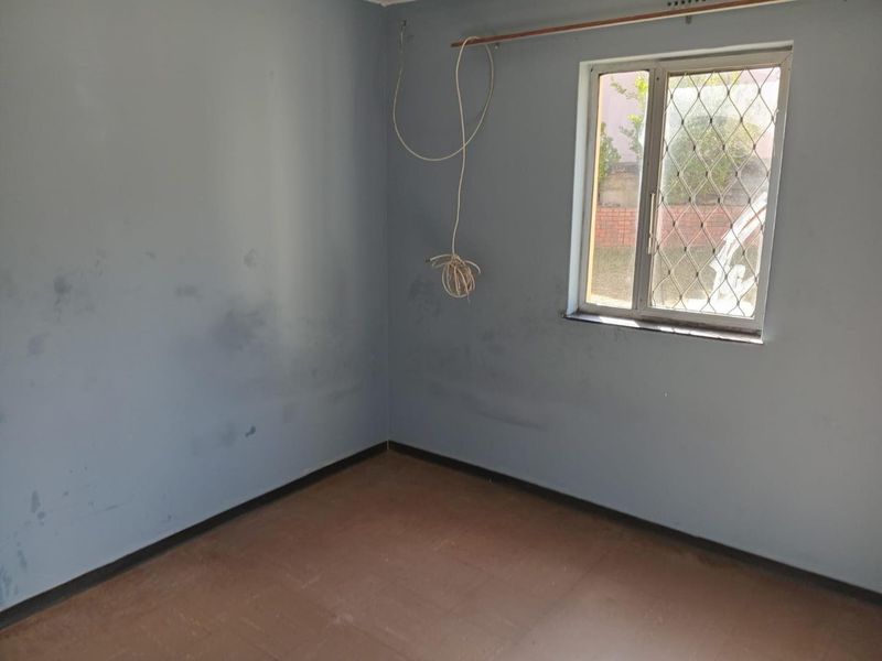 An empty bedroom featuring blue walls, a window with a security gate, and exposed electrical wiring, requiring cosmetic updates and repairs.