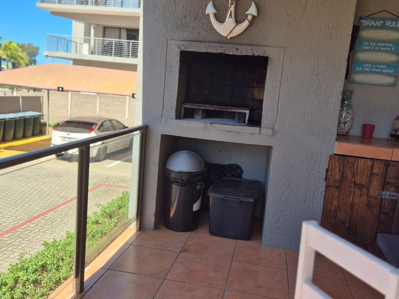 Covered balcony featuring a built-in braai, tiled flooring, and a glass balustrade overlooking the parking area.