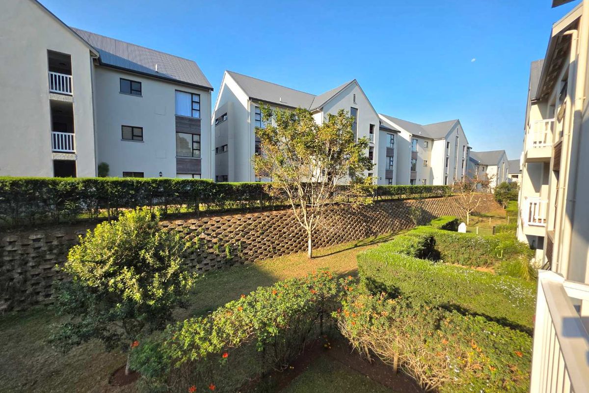 View of the apartment complex exterior, featuring landscaped gardens, manicured hedges, and a retaining wall, viewed from an elevated balcony.