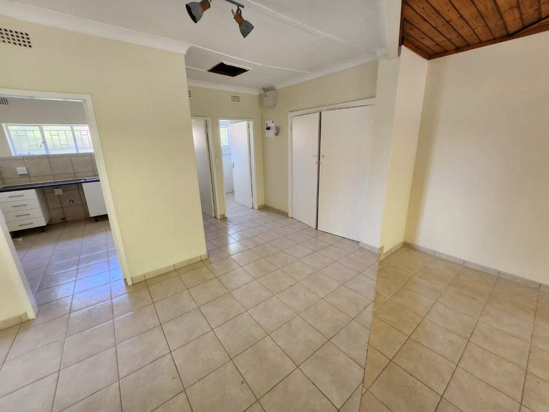 An empty living area featuring tiled flooring, built-in storage cupboards, and a combination of standard and timber-paneled ceilings, leading into a kitchen and hallway.