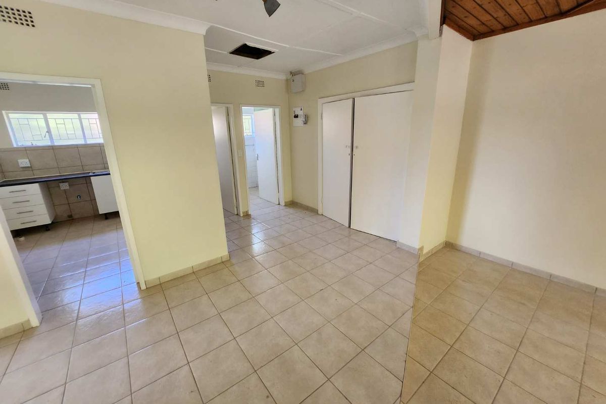 An empty living area featuring tiled flooring, built-in storage cupboards, and a combination of standard and timber-paneled ceilings, leading into a kitchen and hallway.