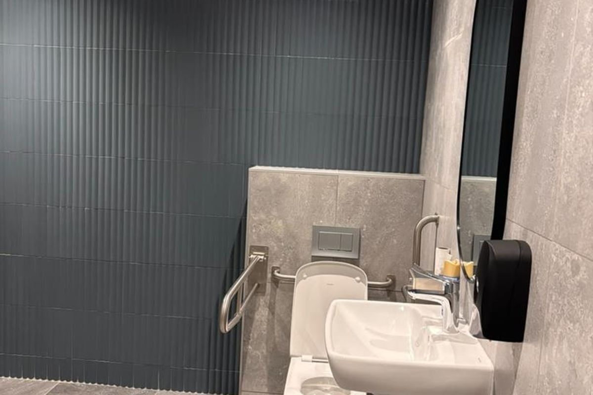 A modern, accessible bathroom featuring high-quality grey tiled finishes, a wall-mounted basin, and a wall-mounted toilet with safety grab rails.