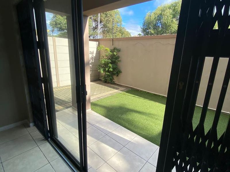 A covered patio area featuring tiled flooring and sliding glass doors, leading out to a private paved and artificial lawn garden space.
