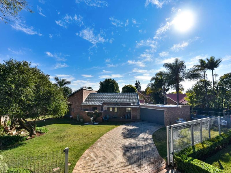 Front view of a brick house featuring a paved driveway, double garage, and a well-maintained garden with mature trees and perimeter fencing.