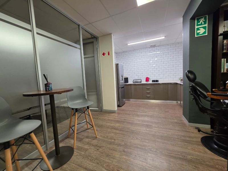 A modern office kitchenette featuring sleek cabinetry, a white subway tile backsplash, and durable laminate flooring, adjacent to a glass-partitioned workspace.