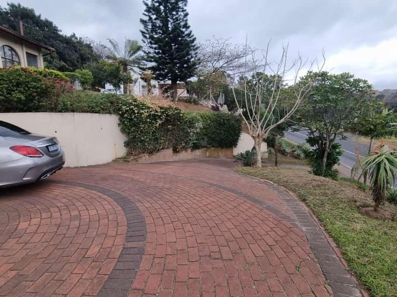 A view of the property's front exterior, featuring a paved circular driveway, a retaining wall, and established garden landscaping.