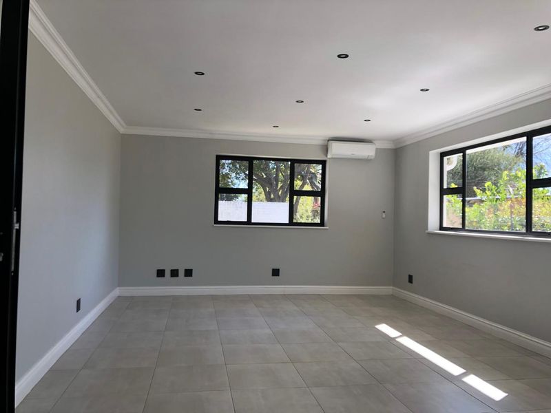 A bright, empty office space featuring neutral grey walls, large black-framed windows, tiled flooring, and a wall-mounted air conditioning unit.