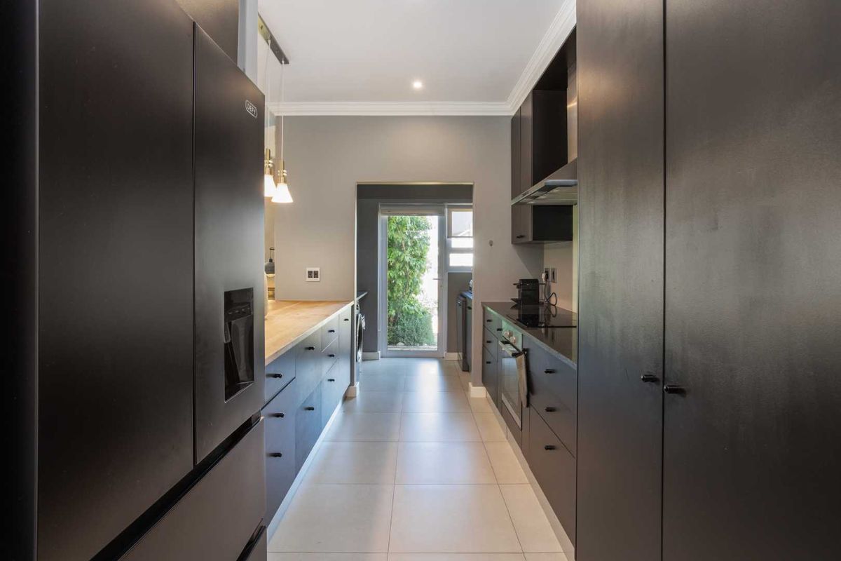 Modern galley-style kitchen featuring sleek dark cabinetry, timber countertops, integrated appliances, and tiled flooring with direct access to the exterior.