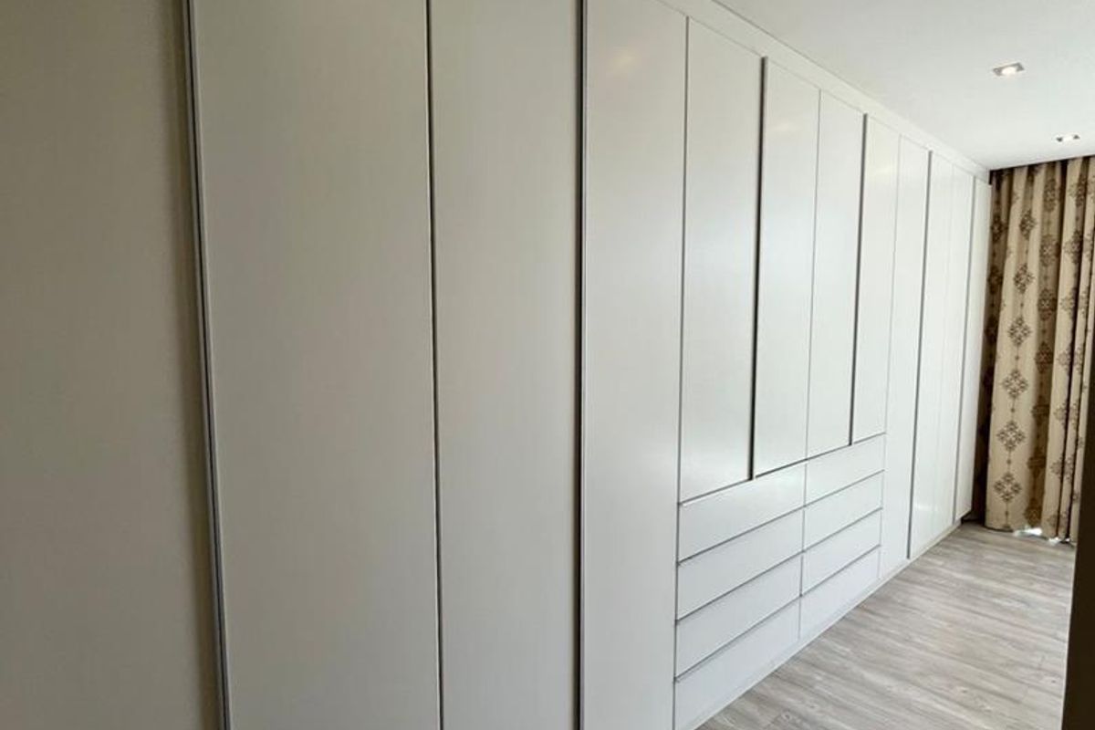 A modern bedroom featuring extensive built-in floor-to-ceiling wardrobes with integrated drawers and sleek laminate flooring.