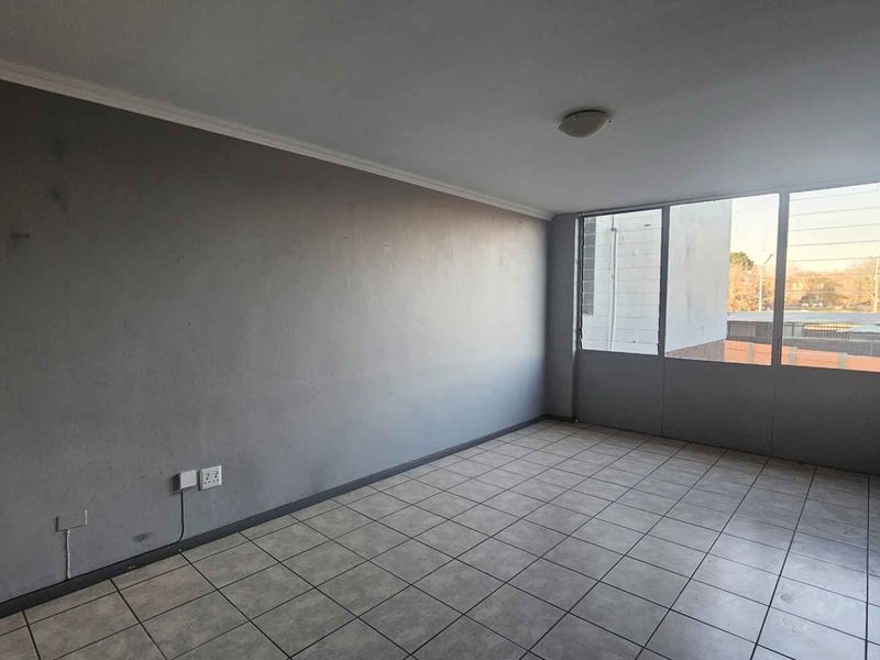 An empty living room featuring tiled flooring, large windows providing natural light, and neutral grey walls in need of minor cosmetic attention.
