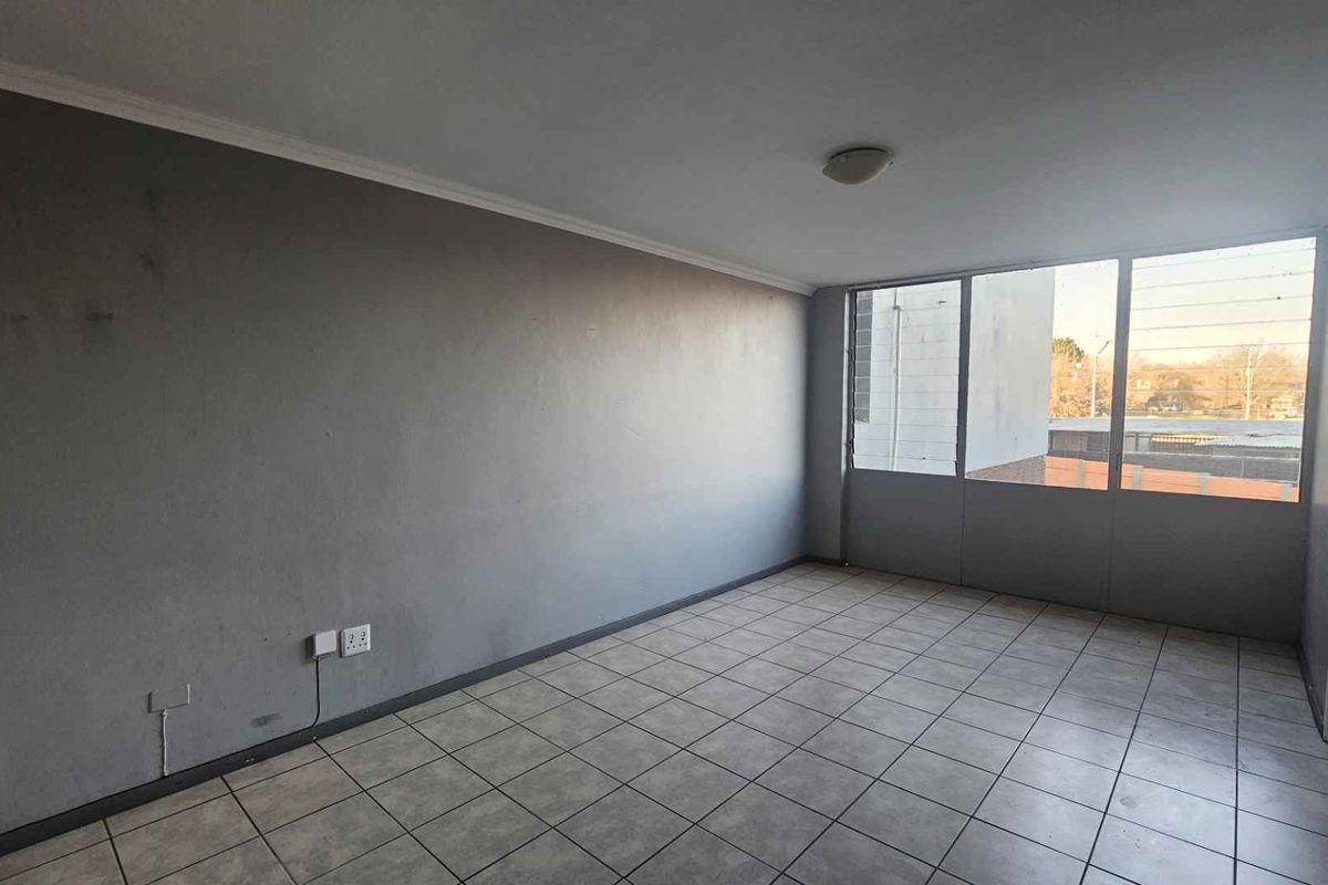 An empty living room featuring tiled flooring, large windows providing natural light, and neutral grey walls in need of minor cosmetic attention.