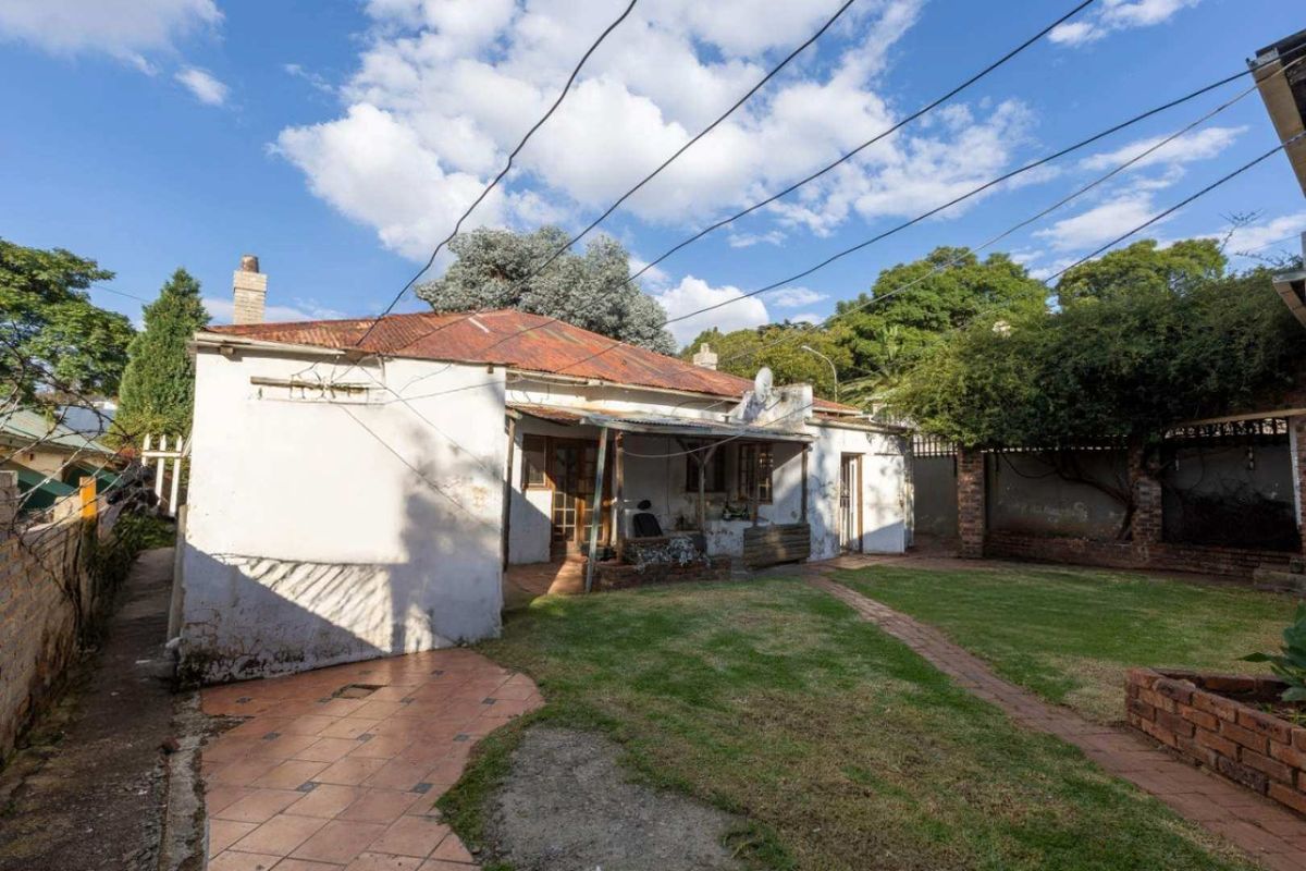 Rear view of the property featuring a garden, paved patio area, and a house with a corrugated iron roof requiring maintenance.
