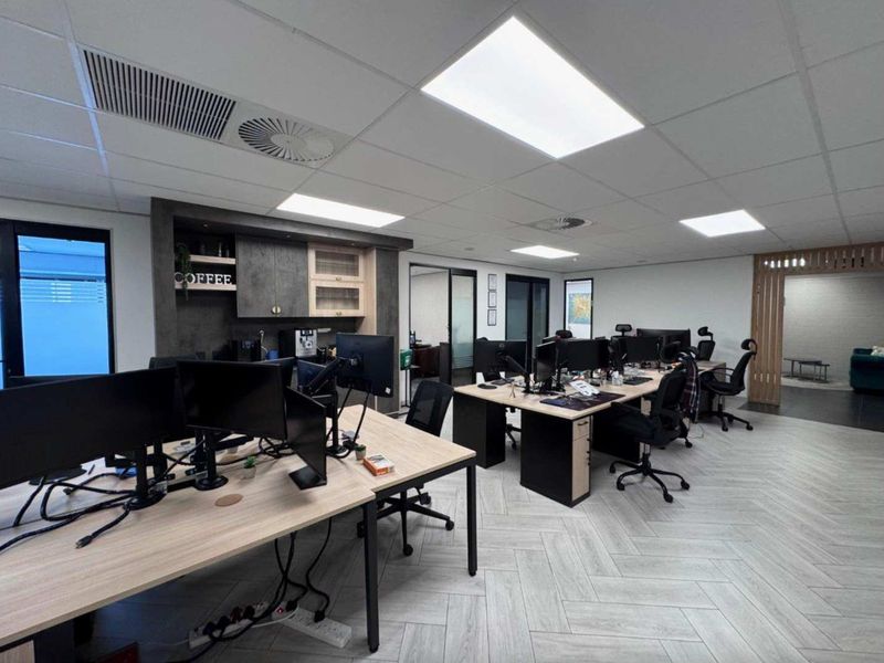 Modern open-plan office space featuring herringbone flooring, suspended ceiling with recessed lighting, integrated kitchenette cabinetry, and glass partition doors.