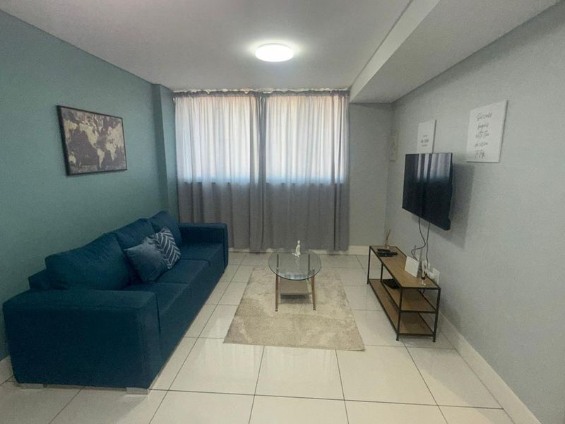 A well-maintained living room featuring neutral tiled flooring, a soft teal accent wall, and large windows with curtains for natural light.